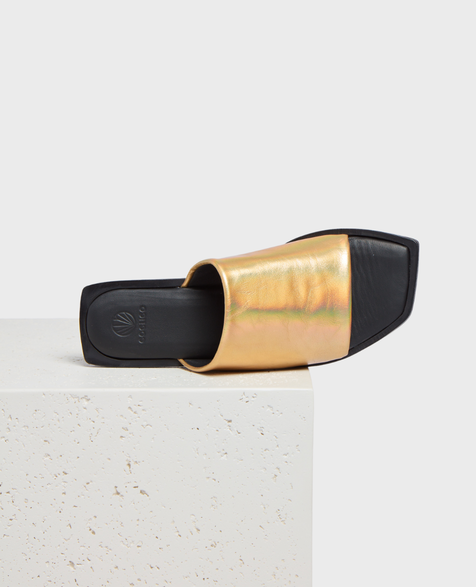 A gold metallic slide sandal with a square toe, black sole, and smooth texture, featuring a wide strap across the top.