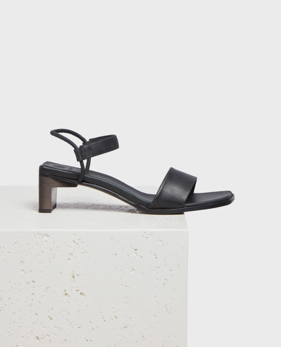 A sleek black sandal with a low block heel, featuring a minimalist design with a single wide strap across the front and a thin ankle strap with a buckle.
