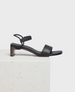 A sleek black sandal with a low block heel, featuring a minimalist design with a single wide strap across the front and a thin ankle strap with a buckle. 1