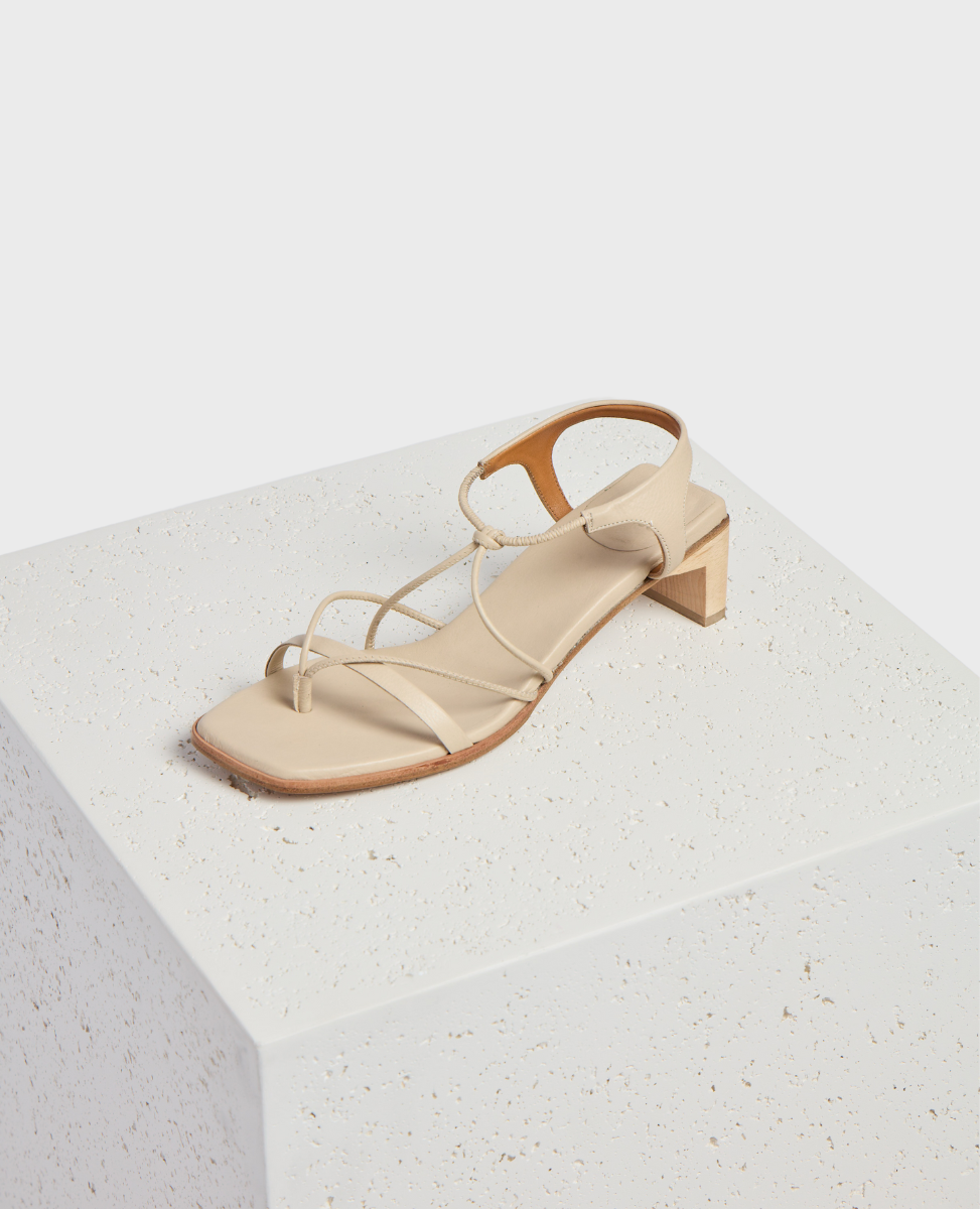 A beige sandal with a square toe, thin crisscrossing straps, a low block heel, and a smooth leather finish rests on a textured white cube.