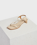 A beige sandal with a square toe, thin crisscrossing straps, a low block heel, and a smooth leather finish rests on a textured white cube. 1