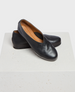 A pair of sleek, black leather slip-on shoes with a smooth texture, rounded toe, and tan interior lining, resting on a textured white surface. 1