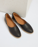 A pair of sleek, black leather slip-on shoes with a smooth texture, almond-shaped toe, tan interior lining, and a light brown flat sole. 1