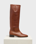 A tall, sleek brown leather boot with a smooth, polished finish, featuring a rounded toe, low block heel, and subtle stitching details, standing on a white platform. 1