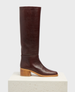 A tall, sleek, dark brown leather boot with a smooth texture, featuring a rounded toe and a sturdy, light wooden block heel, standing on a textured white platform. 1