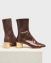 The product is a pair of metallic brown ankle boots with a sleek, pointed toe, featuring a smooth, shiny texture, a side zipper, and a distinctive light wooden block heel. 2