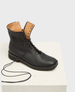 A sleek black leather boot with a smooth texture, featuring a high ankle design, a side zipper, and tan interior lining, resting on a white textured platform. 3