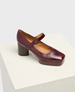 A burgundy Mary Jane shoe with a glossy leather finish features a chunky block heel, a rounded toe, and an adjustable strap with a buckle, set on a textured white platform. 2