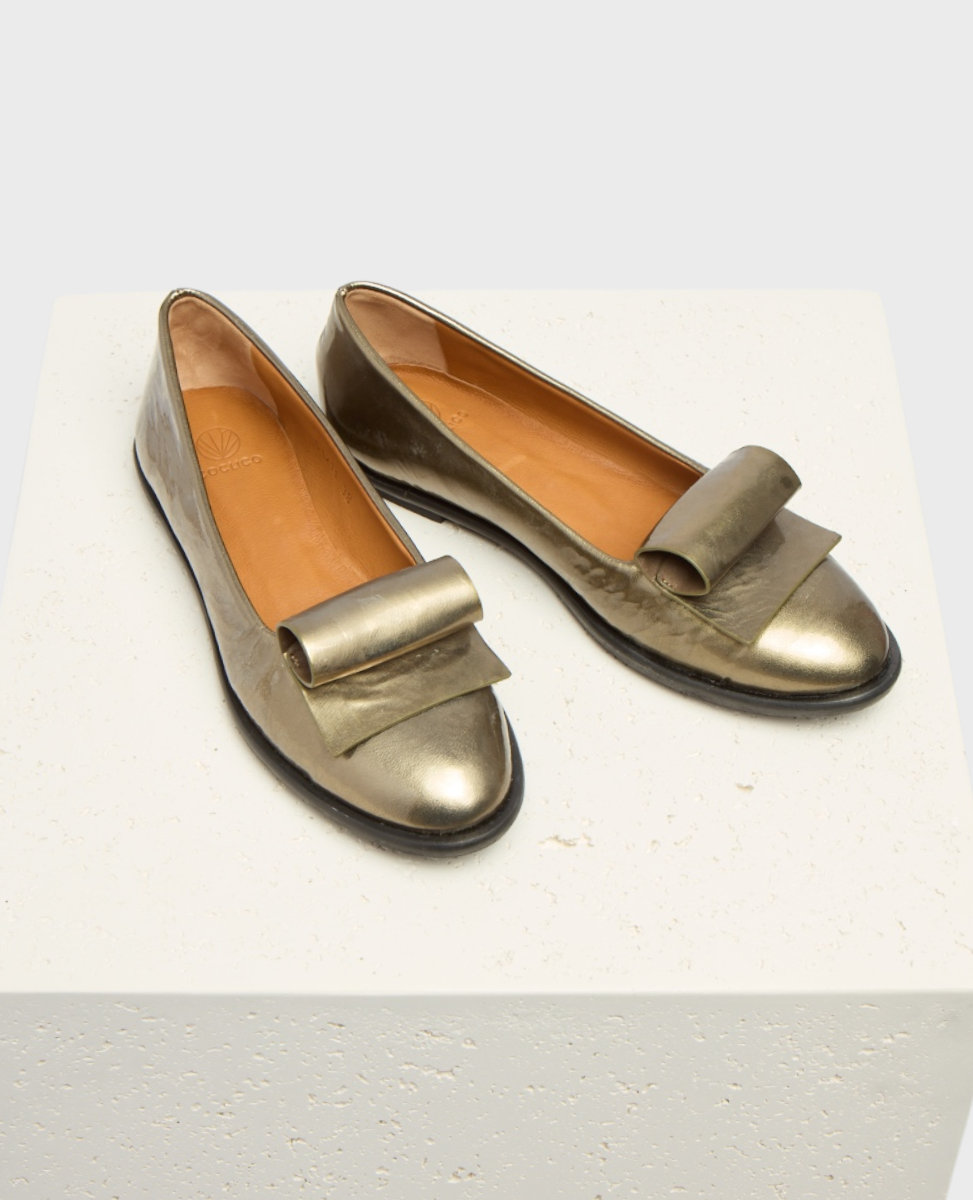 Metallic bronze flat shoes with a smooth, shiny texture, featuring a distinctive wide bow on the front, a rounded toe, and a tan interior lining.