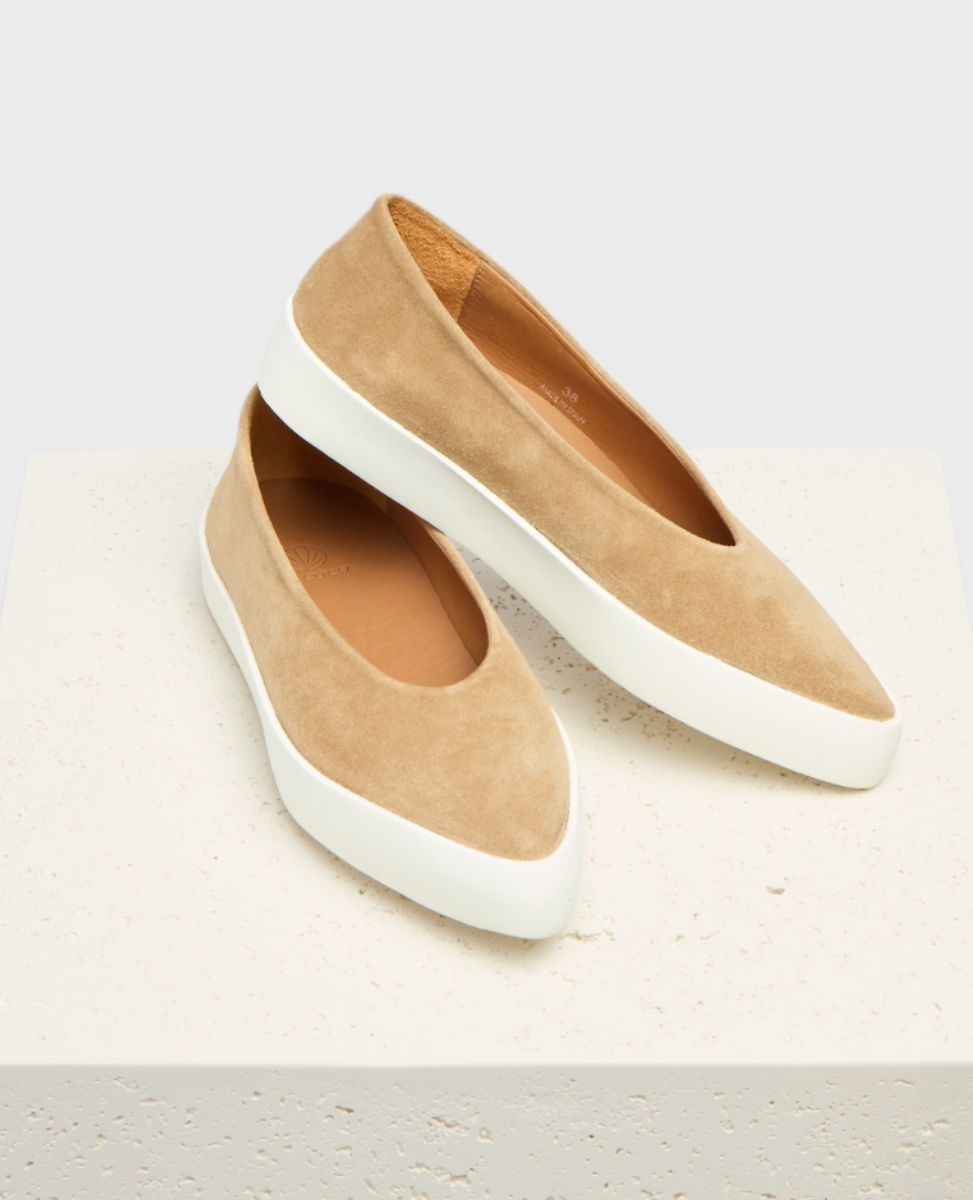 A pair of beige suede slip-on shoes with a smooth texture, featuring a sleek, rounded design and contrasting thick white rubber soles.