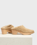 A pair of tan suede clogs with a plush, fur-lined interior, featuring a chunky wooden heel and platform, showcasing a smooth, rounded toe design. 1