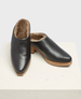 A pair of sleek, black leather slip-on clogs with a smooth finish, featuring a plush brown fur lining, a slightly pointed toe, and a light brown wooden sole with a low heel. 2