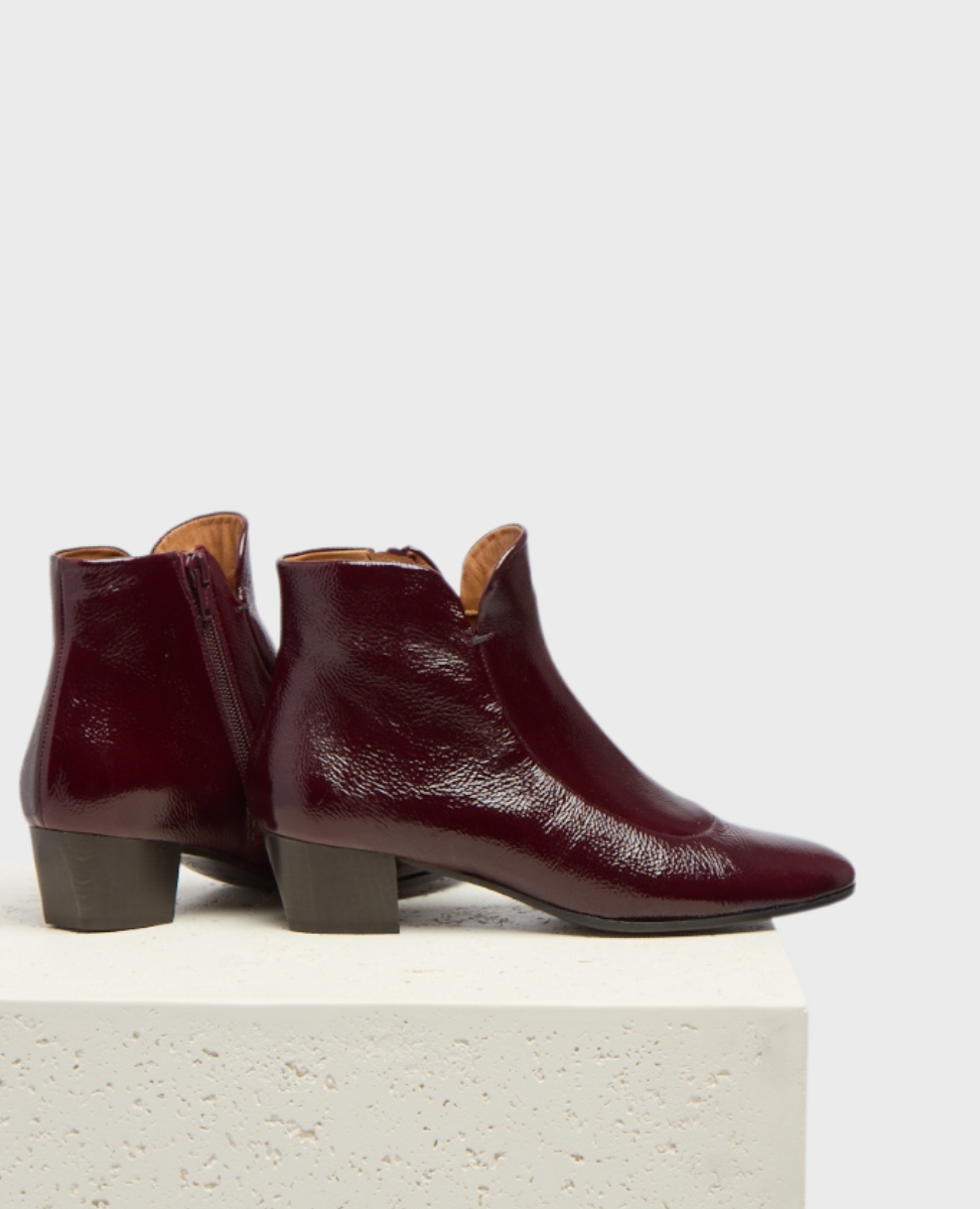These ankle boots feature a glossy burgundy leather exterior with a sleek, rounded toe, a low block heel, and a side zipper for easy access.