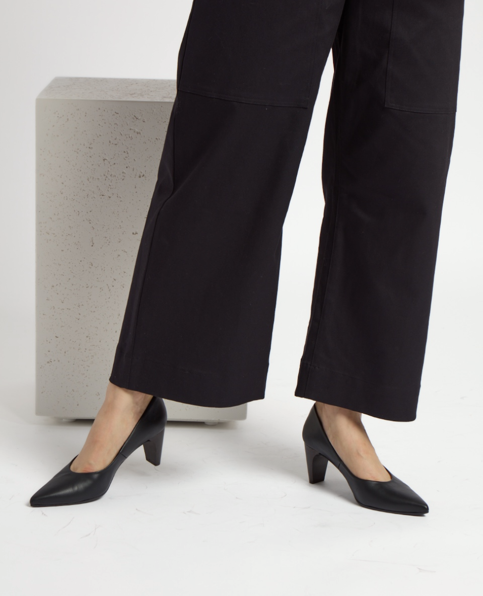 A pair of sleek, black leather pumps with a pointed toe and a mid-height wood heel, worn by a person in black wide-leg pants, standing on a smooth, light gray surface.
