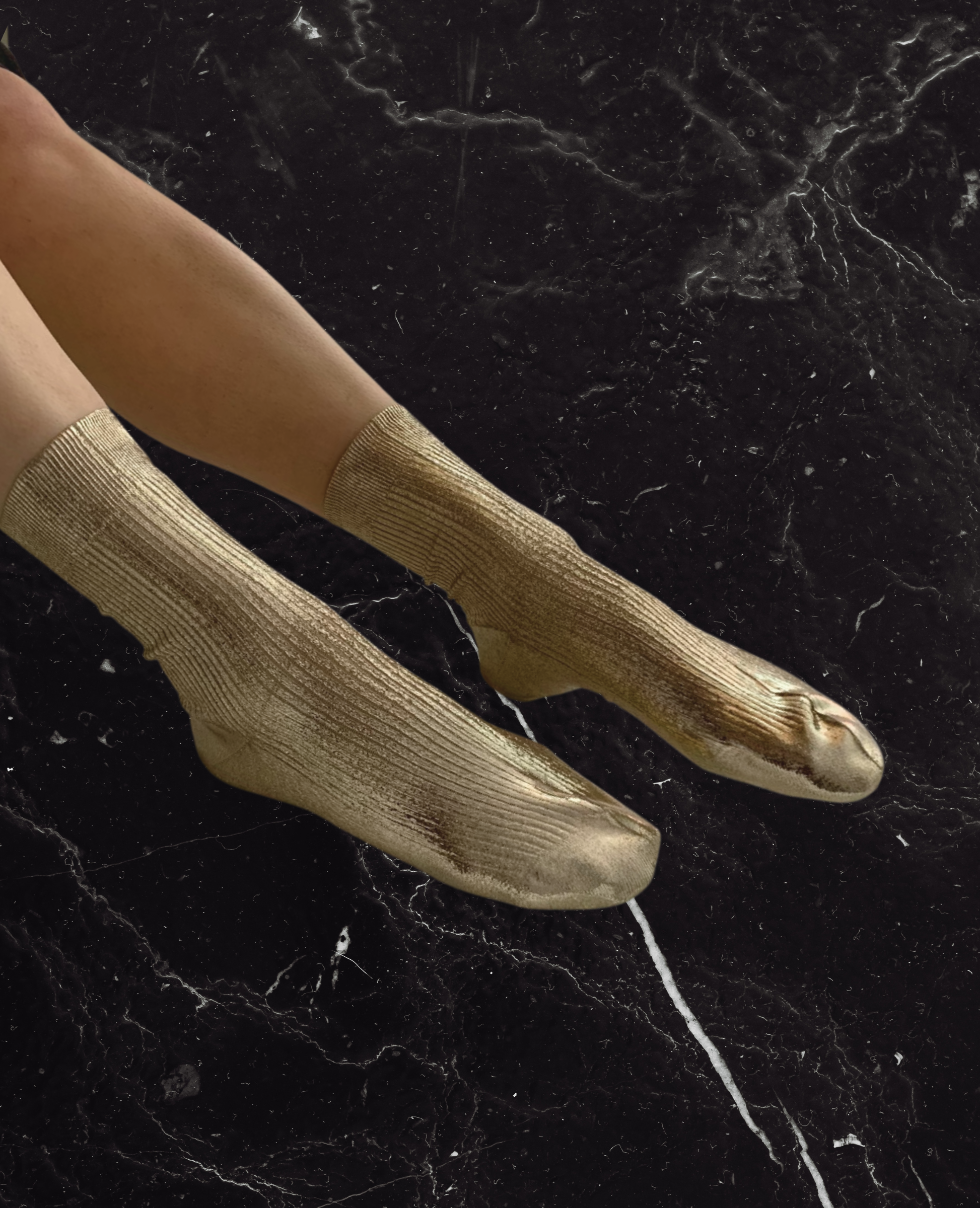 Gold metallic socks with a ribbed texture, mid-calf length, and a slightly shiny finish, worn on a pair of feet against a dark, marbled background.