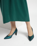 A pair of elegant, teal-colored high-heeled shoes with a pointed toe design, smooth leather texture, and a medium-height block heel, worn with a matching green dress. 4