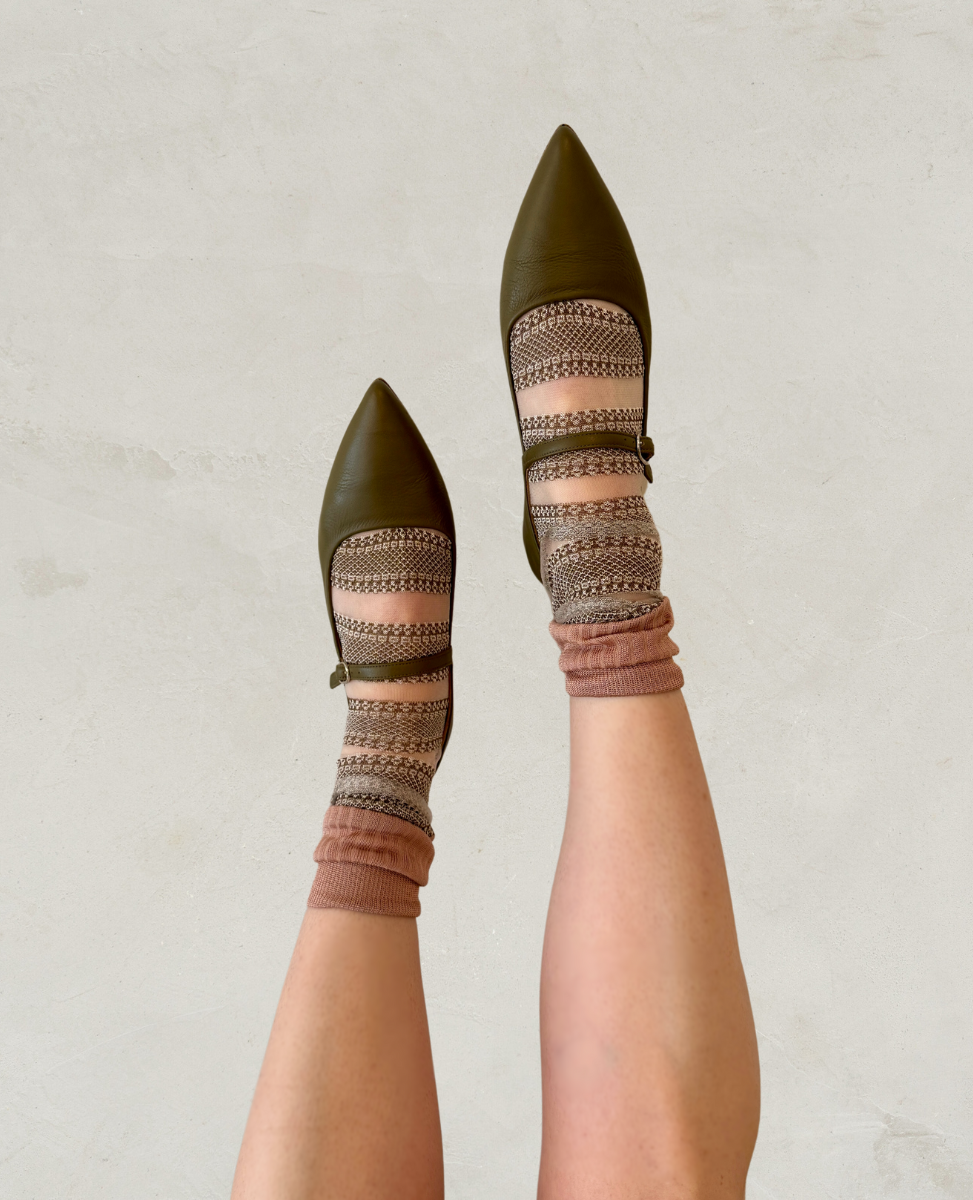 Pointed-toe olive green flats with a smooth leather finish feature a strap across the top, paired with patterned socks in shades of brown and beige.