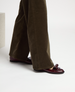 A pair of burgundy leather loafers with a glossy finish, featuring a rounded toe and a decorative strap across the top, worn with brown corduroy pants. 3