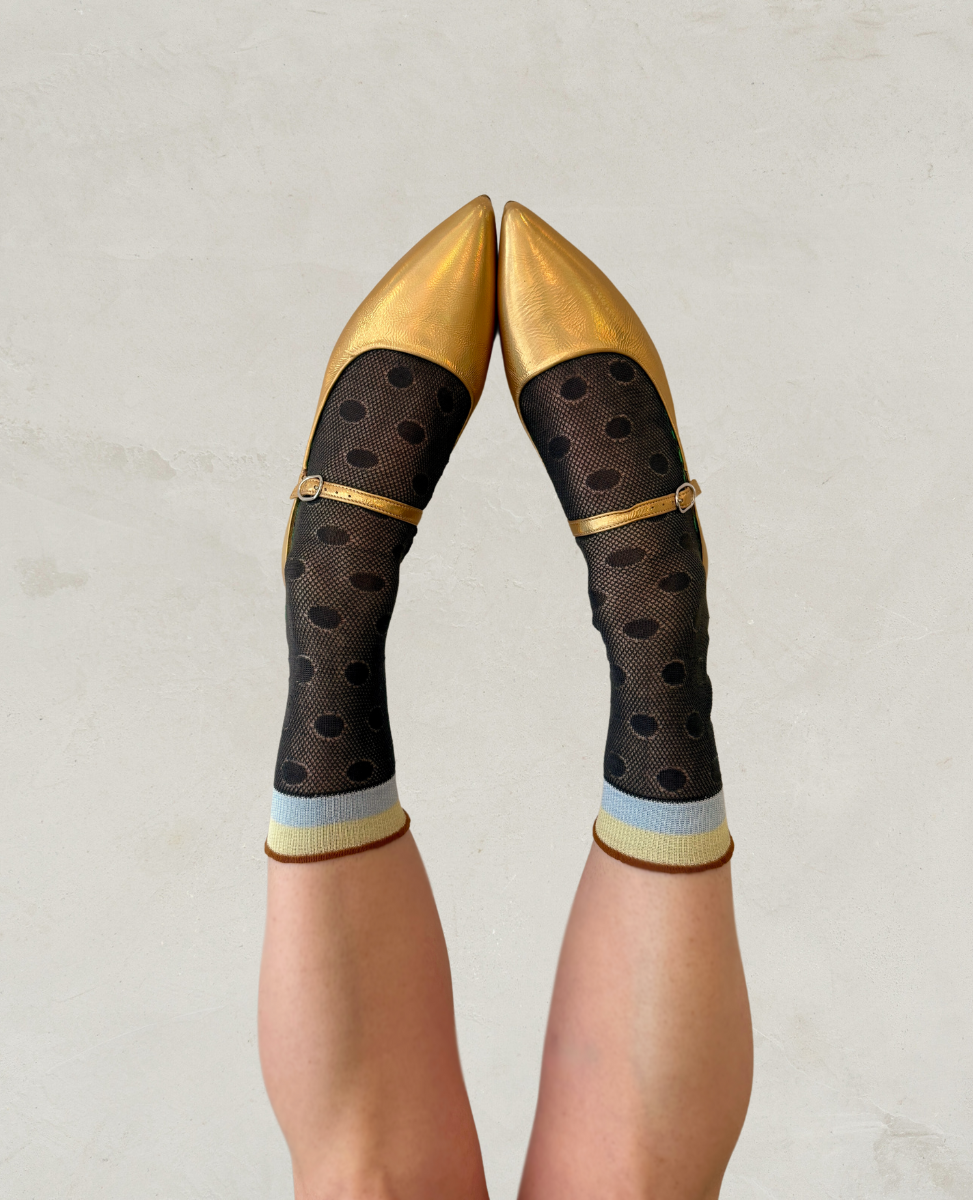 A pair of pointed, metallic gold shoes with a strap across the top, worn over black mesh socks featuring large polka dots and light blue cuffs.