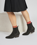 Black ankle boots with a pointed toe, low block heel, elastic side panels, and a pull tab at the back, worn with red socks and a black skirt. 2