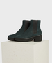 Dark green suede ankle boots with a chunky black heel, featuring elastic side panels and a textured sole, resting on a light-colored textured block. 2