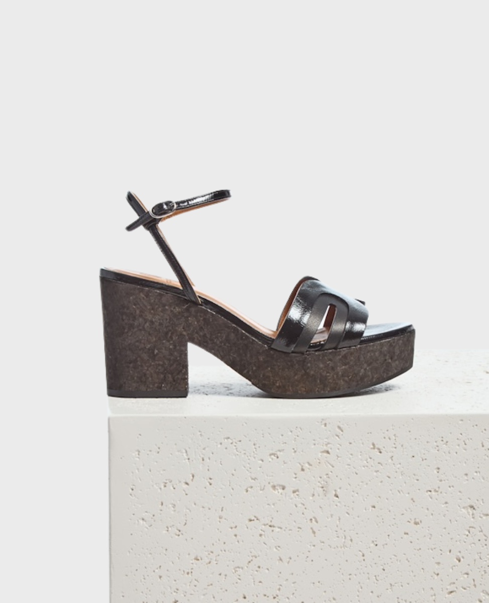 A black sandal with a glossy finish features a thick cork platform heel, an open toe design with two wide straps, and a thin ankle strap with a buckle closure.