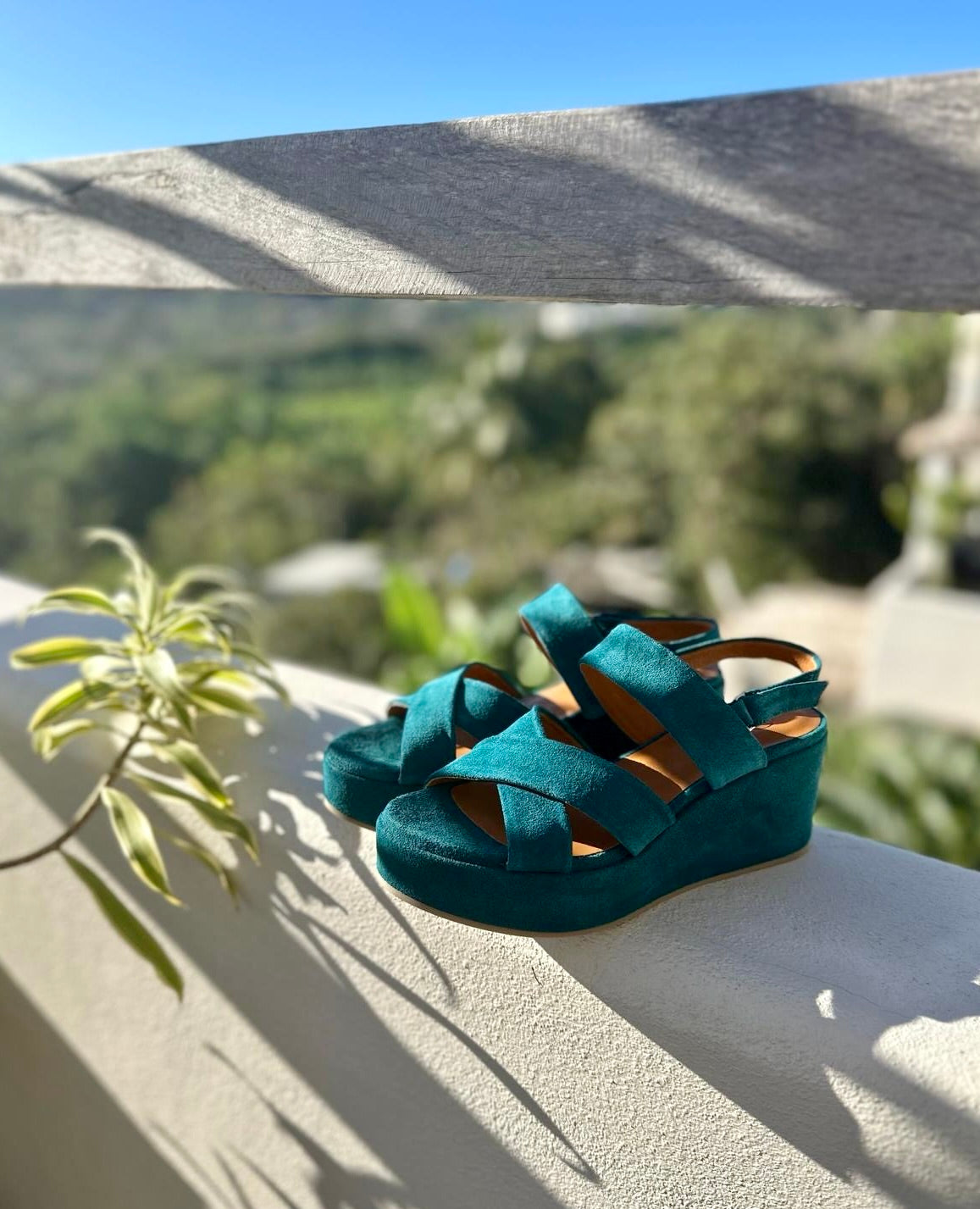 Lacine Wedge sandal in Azure split suede, on a wall with trees in the background