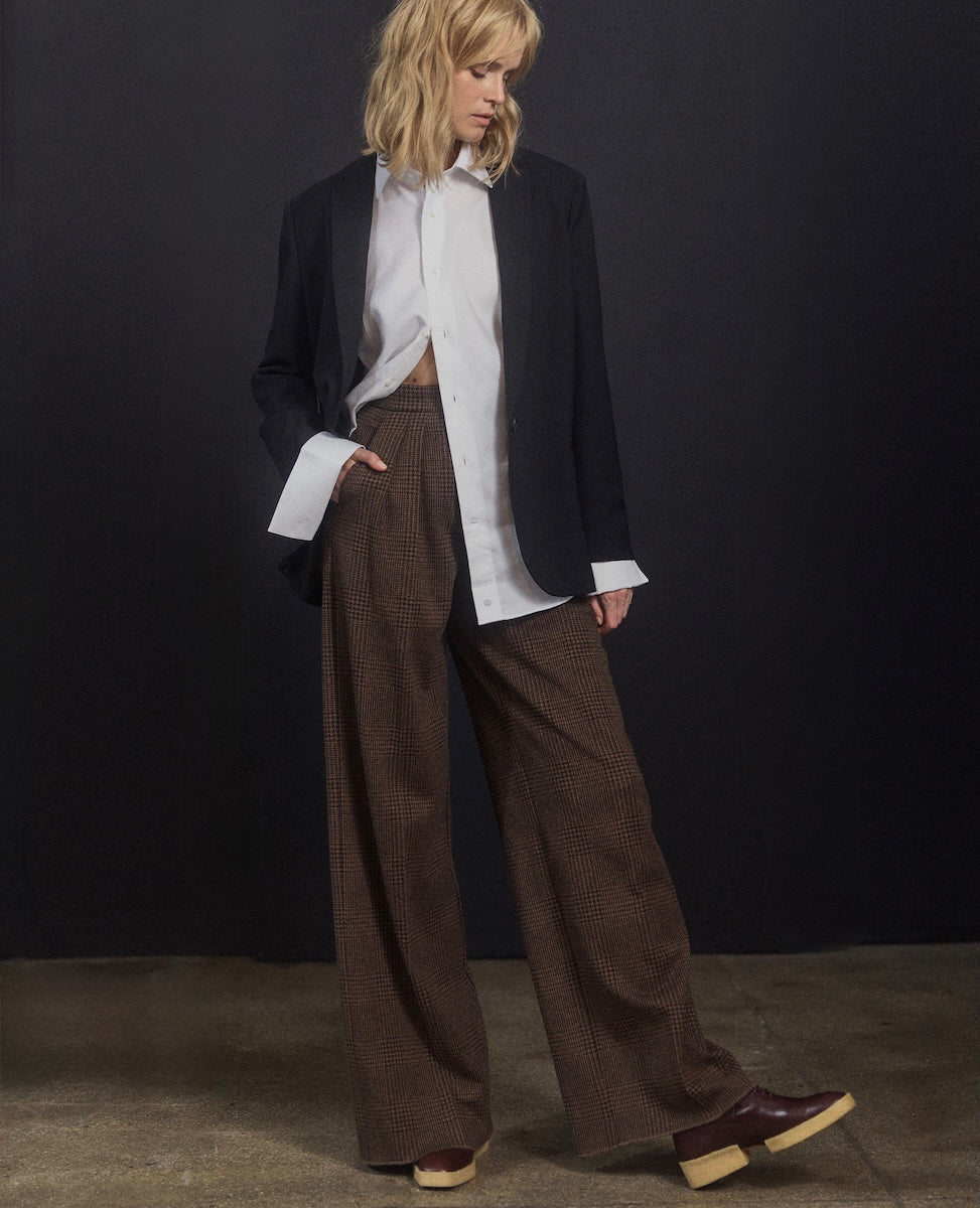 Person wearing a black blazer, white shirt, and brown checkered pants against a dark background