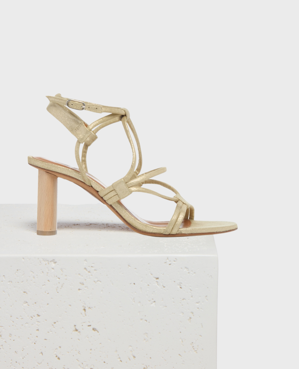 A gold strappy sandal with a block heel made of light wood, featuring a delicate ankle strap and a minimalist design.