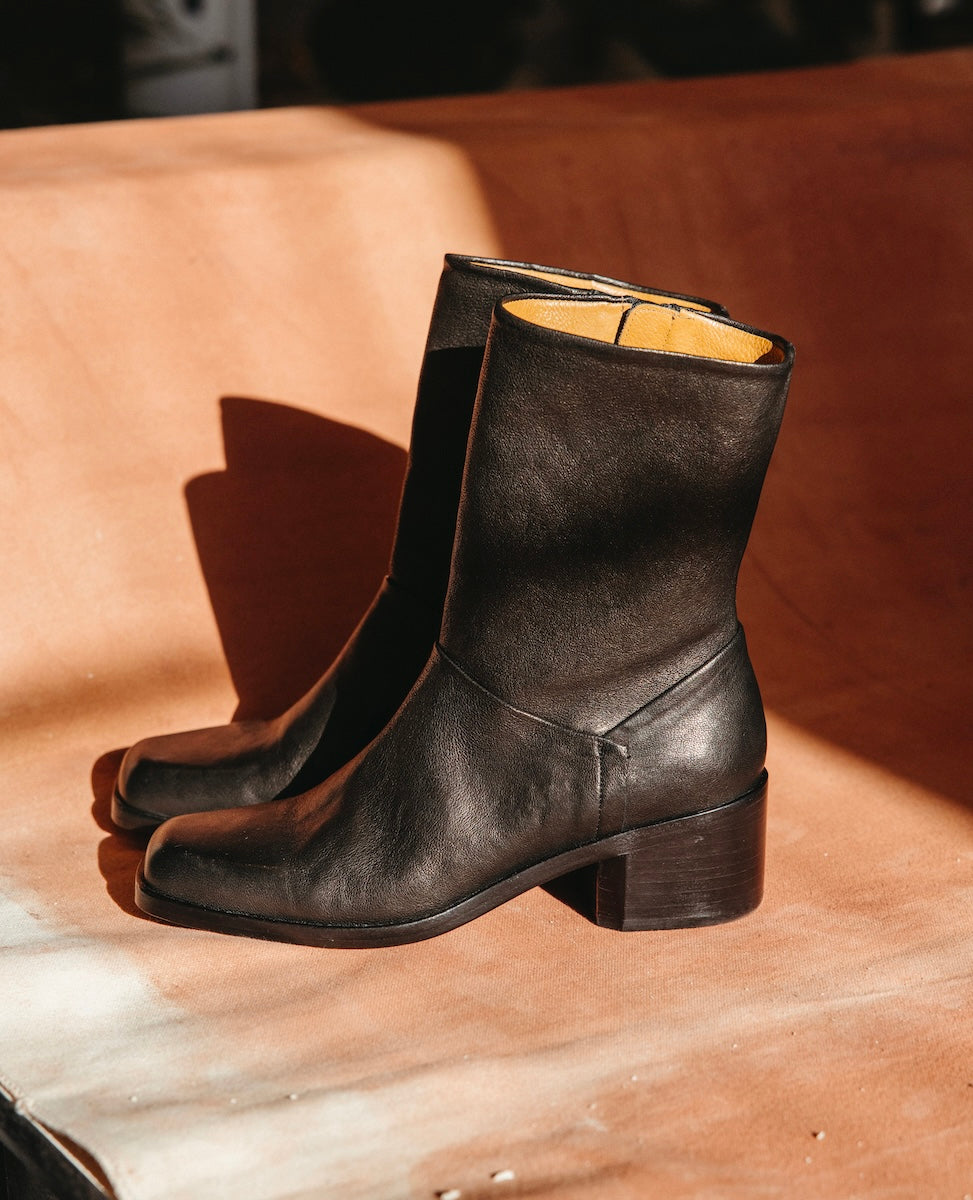 A pair of mid-calf black leather boots with a smooth texture, a square toe, a sturdy stacked  heel, and a tan interior lining.