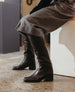 A pair of sleek, black leather knee-high boots with a smooth texture, a modern square toe, and a mid-height stacked heel, worn with a gray wool skirt. 10