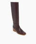 A tall, brown leather boot with a smooth texture, a slightly rounded toe, a low wooden block heel, and a sleek, minimalist design. 3