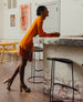 Woman wearing an orange fringed dress smiling and leaning against a bar, wearing the Angel Heel in grey.  5