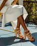 Lower half of a woman sitting down on a yellow chair, wearing a white skirt and the April Heel in Fawn.  4