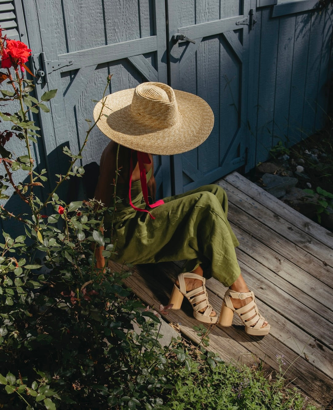 Woman sitting next to a garden wearing a straw hat, green jumpsuit and the April Heel in Latte Machiatto. 