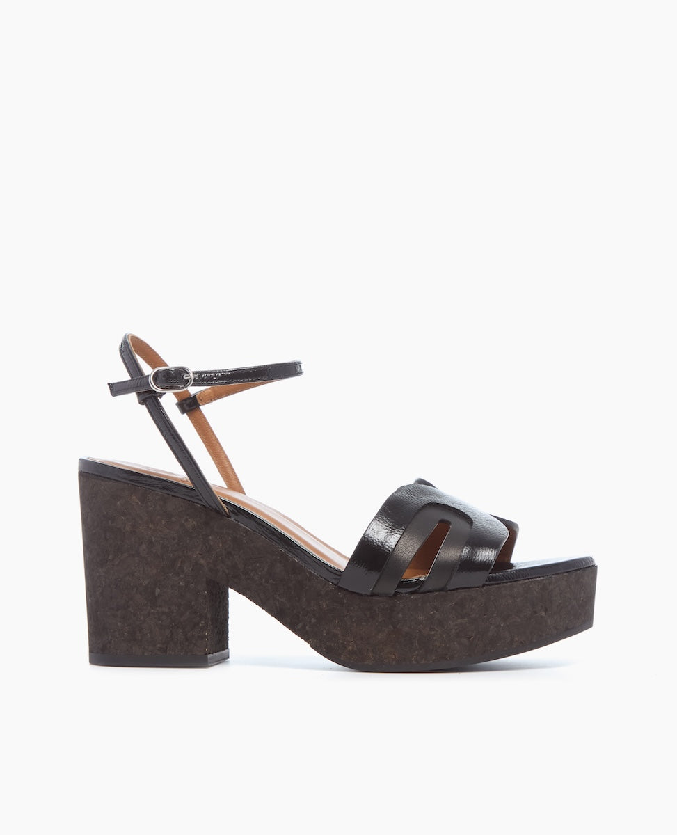 A black sandal with a chunky cork heel, featuring a strappy leather upper and an adjustable ankle strap with a small buckle.