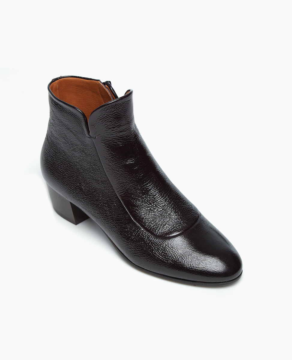 The Calm Boot in amethyst patent: classic black leather grounded by a solid wood angled heel, featuring a bid front insert and an almond toe shape. Angled view.