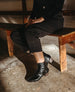 A person is seated on a wooden bench, wearing black ankle boots with a glossy finish, an almond shaped toe, and a low, solid wood heel, paired with black pants. 9