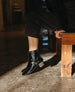 A pair of black, shiny, ankle-high boots with a slight heel, worn by a person dressed in black pants, standing on a polished concrete floor next to a wooden bench. 10