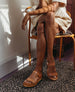 Woman sitting on a wooden chair with her arms atop her crossed legs wearing the Dana Wedge in Savana Cuoio. 3