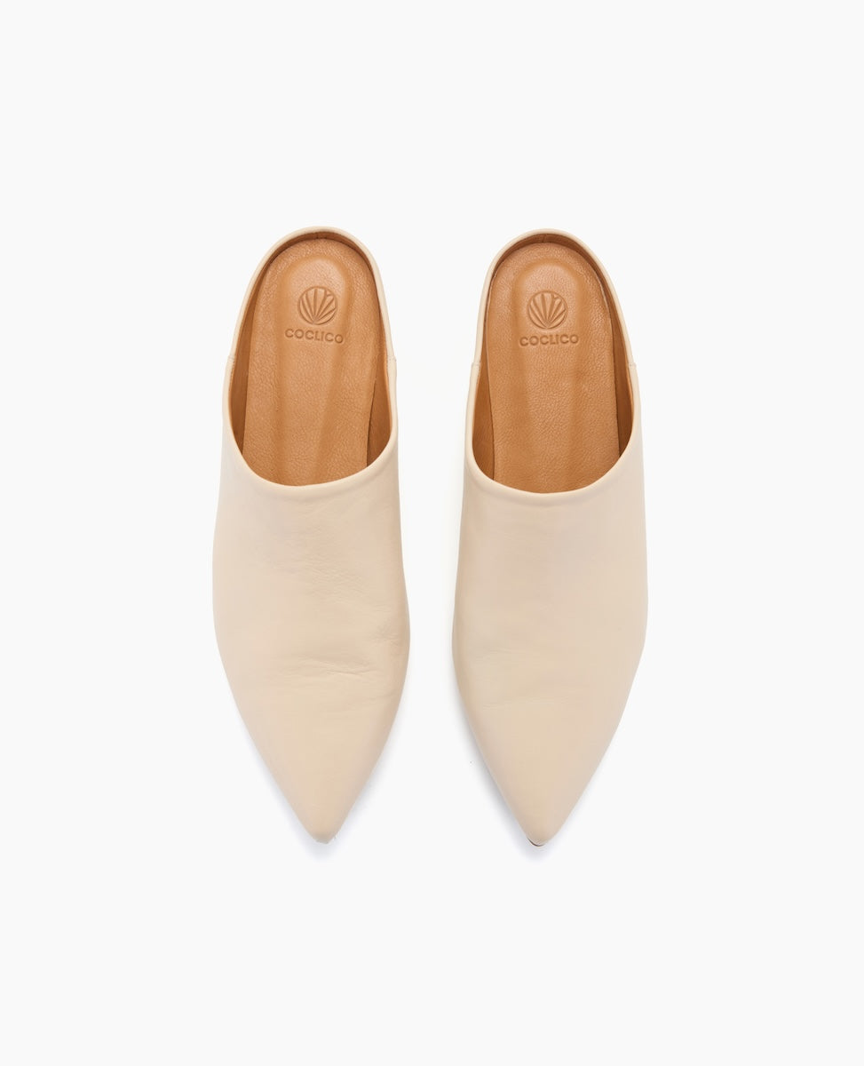 A pair of sleek, cream-colored, pointed-toe mules with a smooth leather texture and tan insoles featuring a subtle logo imprint.
