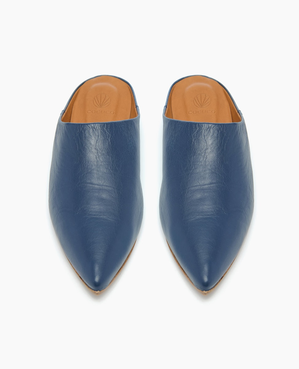 A pair of sleek, pointed-toe mules in a rich blue leather with a smooth texture, featuring a tan insole and a minimalist design.