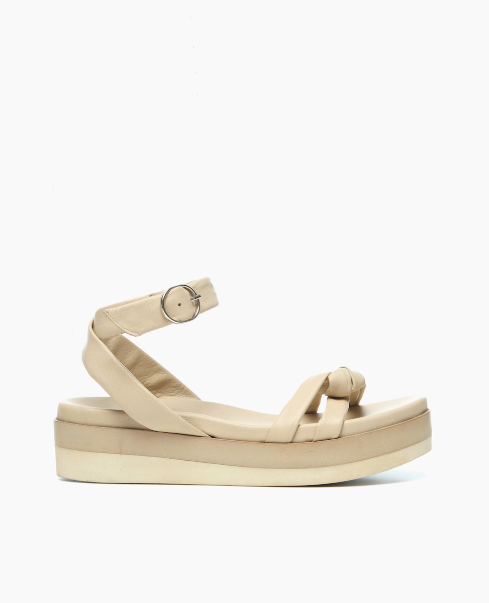 A beige sandal with a thick platform sole, featuring a crisscross strap design and an ankle strap with a silver buckle, made from smooth leather material.