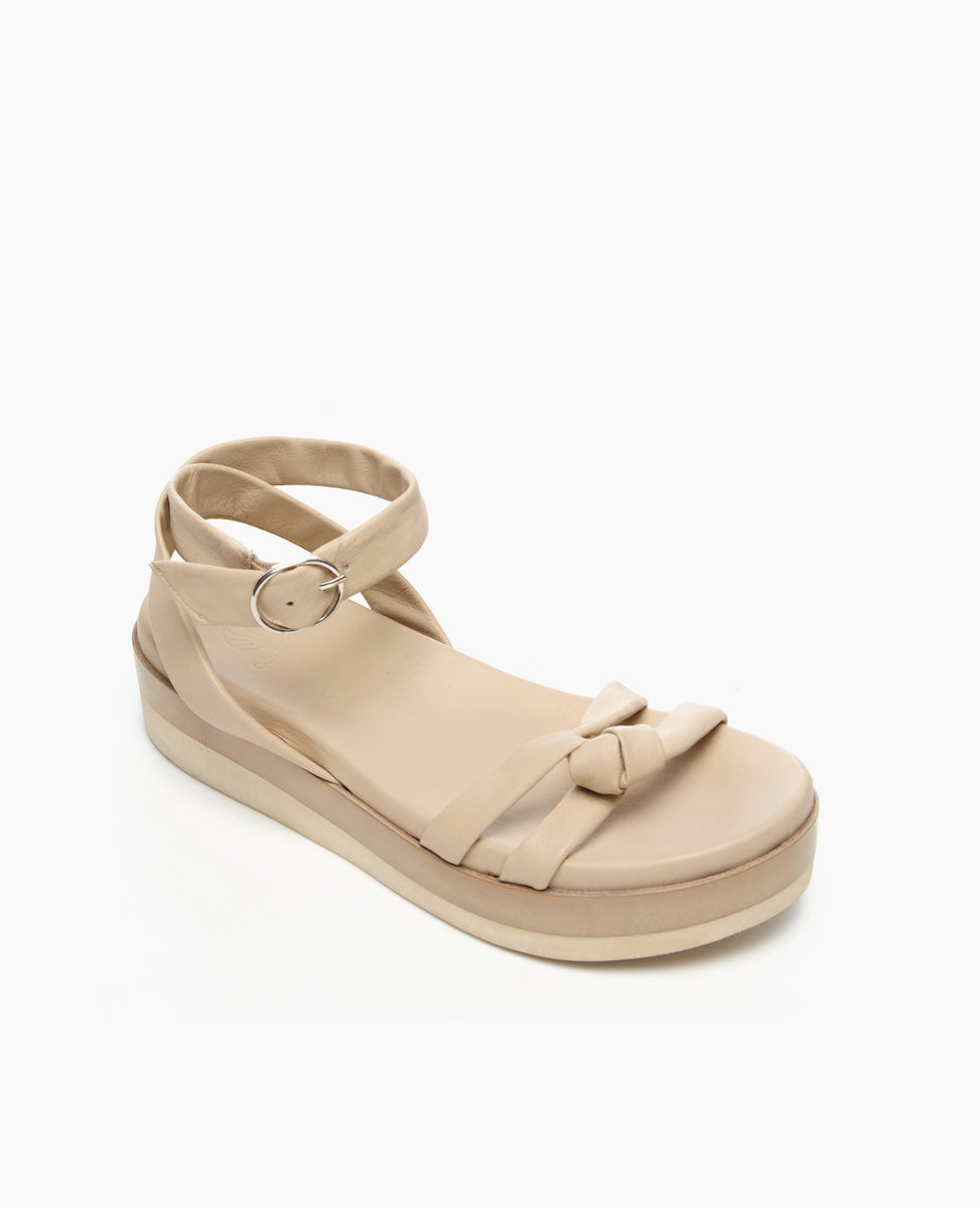 A beige sandal with a flat platform sole features a smooth texture, an ankle strap with a silver buckle, and a decorative knot on the front strap.