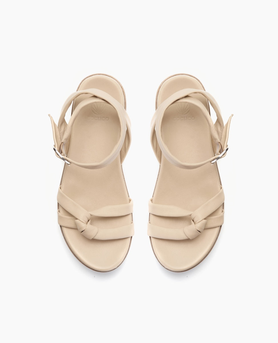 Beige sandals with a smooth texture feature two knotted straps across the toes, an adjustable ankle strap with a silver buckle, and a flat sole.
