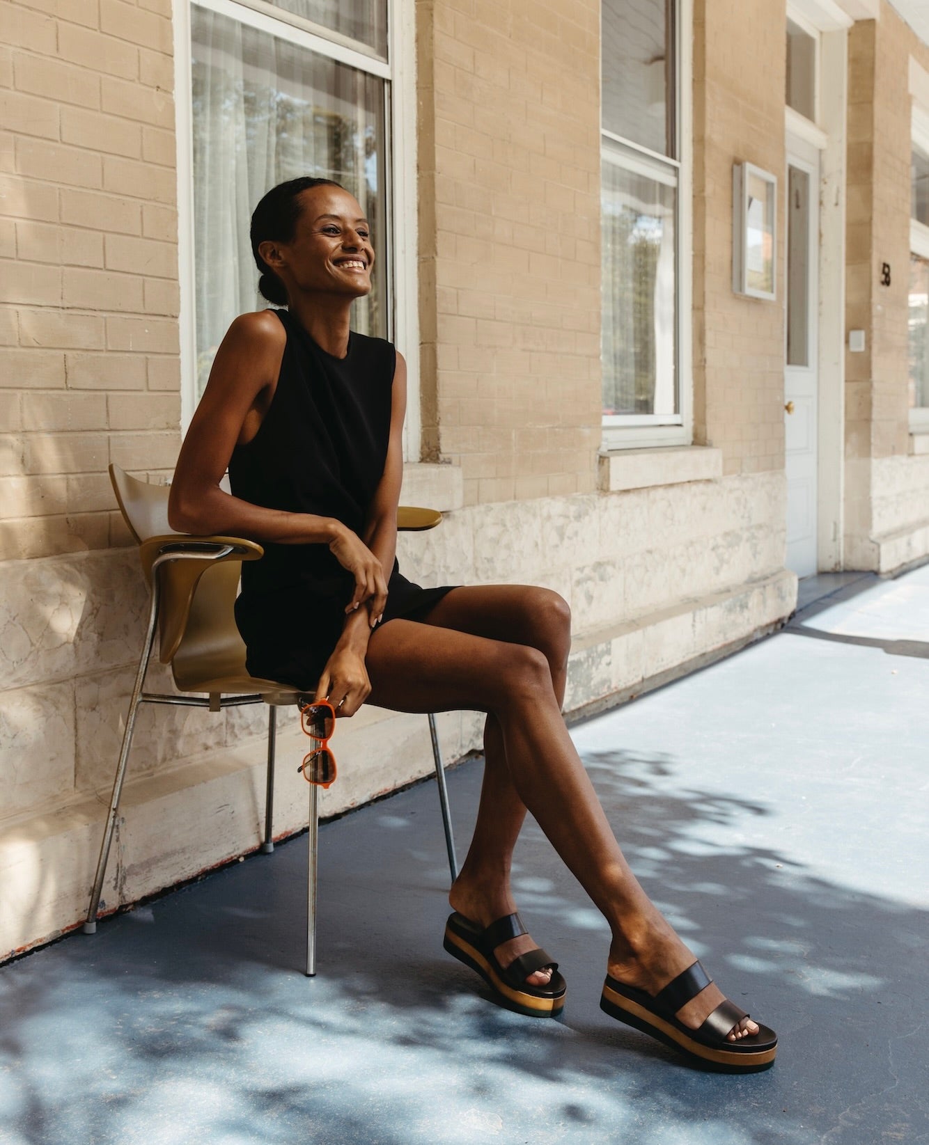 Woman smiling in a black dress, sitting on a wooden chair  wearing the Dana Wedge in Black. 