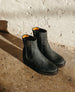 A pair of black leather Chelsea boots featuring elastic side panels, a rounded toe, a mid-height platform heel and a contrasting tan interior lining, set against a textured concrete floor and a rough white wall. 8