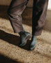 A pair of deep sea split suede ankle boots with black elastic side panels, a rounded toe, and a mid-height platform heel, worn with dark brown pants. 10