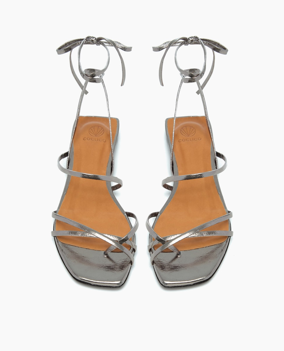 Silver high-heeled sandals with a square toe, featuring thin metallic straps that wrap around the foot and ankle, and a tan insole with a subtle logo imprint.