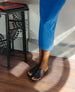 Legs and feet of a women wearing a blue dress and the Ferhana Sandal in anthracite shimmer suede to the right of a black side table.  7